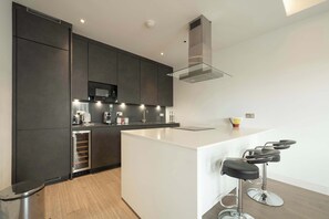 A modern kitchen with dark cabinets, a stainless steel range hood, and a white island with bar stools. - Beautiful, modern, light flat in Shoreditch (a) (Greater London)