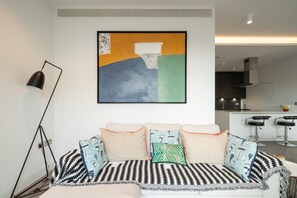 Una sala moderna con un sofá, cojines, una lámpara de pie y una cocina al fondo.