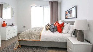Ein modernes Schlafzimmer mit Bett, Nachttisch, Lampe und einem großen Fenster mit Vorhängen.