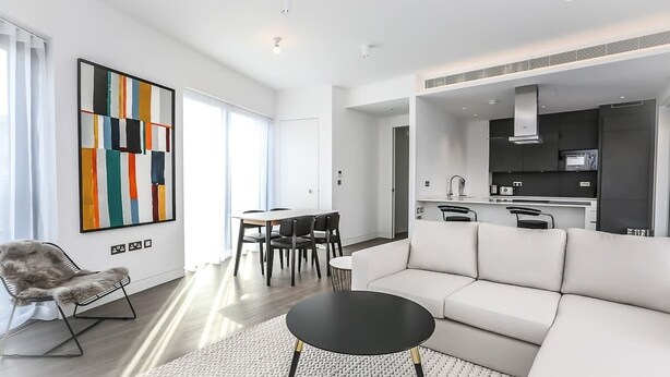 A modern living room with a white sofa, a black coffee table, and a dining area with a table and chairs. There is a kitchen area with a microwave and stove in the background.