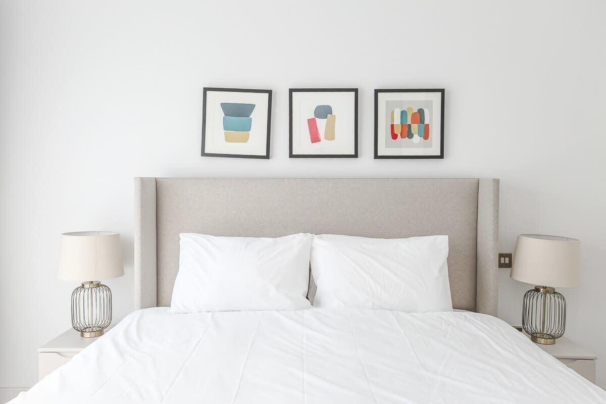 A neatly made bed with white bedding, a grey headboard, and two bedside lamps with a wireframe design. Three framed artworks are hung above the bed.
