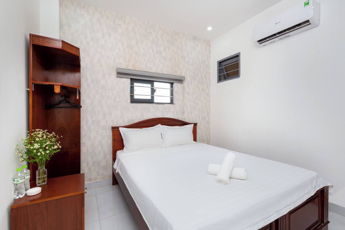 A neatly made bed with white linens, a wooden headboard, a small window with blinds, and an air conditioning unit on the wall.