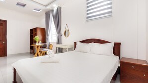 Une chambre d’hôtel avec un grand lit, une table de chevet en bois, un bureau avec une chaise, une petite table avec une plante et une fenêtre avec des stores.
