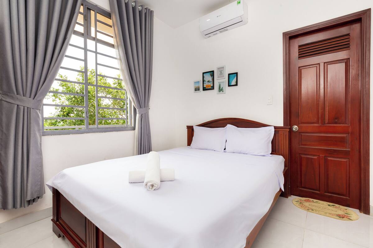 A neatly made bed with white linens, a wooden headboard, and a window with grey curtains.