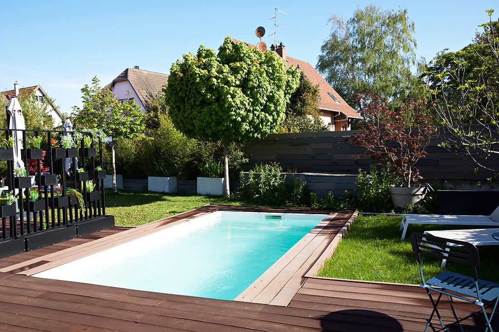 A rectangular swimming pool surrounded by a wooden deck, with a chair and umbrella on one side.