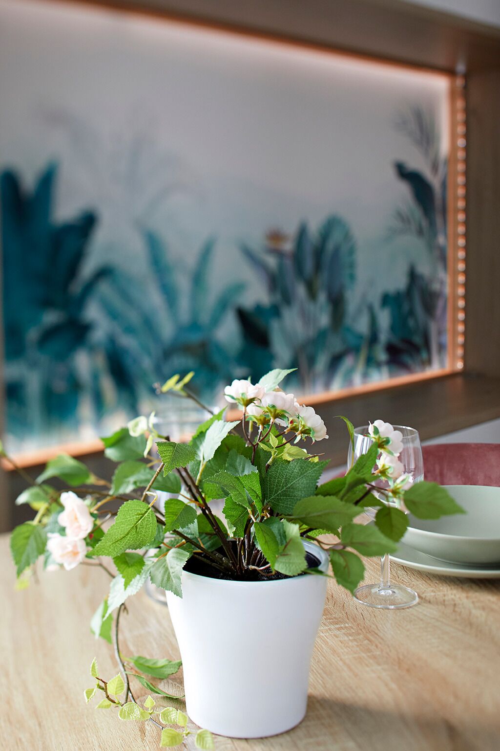 Uma planta branca em vaso com flores brancas sobre uma mesa de madeira, com uma pintura ao fundo.