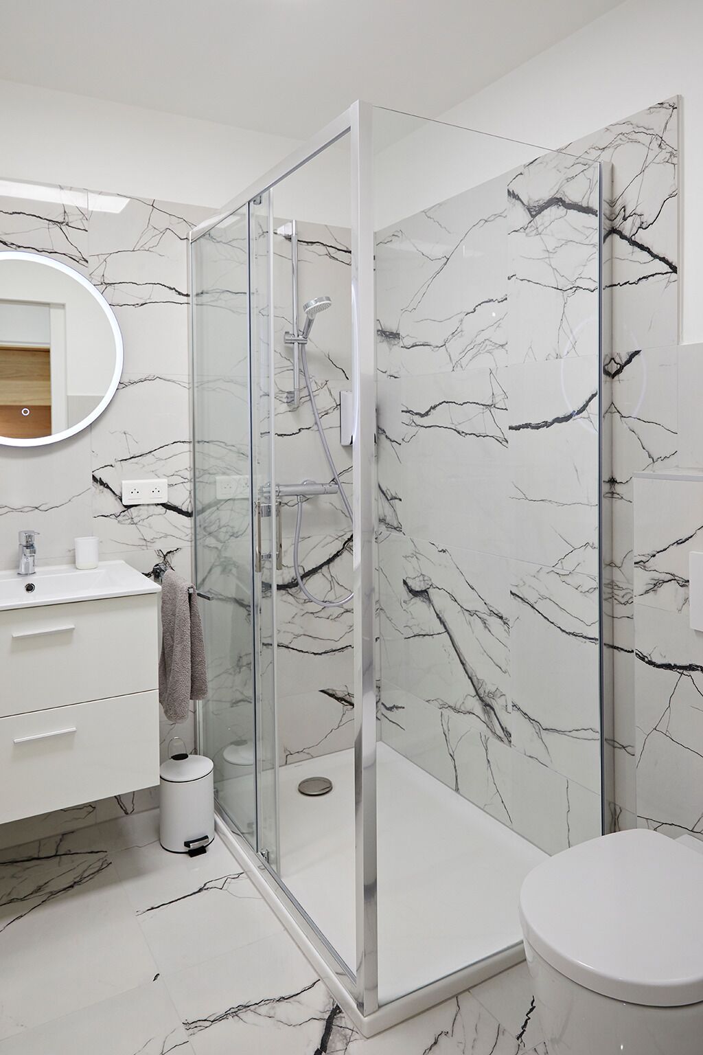 Een moderne badkamer met een douchecabine van glas, wanden met marmerprint, een witte wastafel en een ronde spiegel.