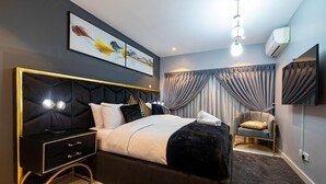 A modern bedroom with a black upholstered bed, a gold-framed nightstand, a grey armchair, and a large window with grey curtains. - EBS LEGEND GHANA  (Accra)