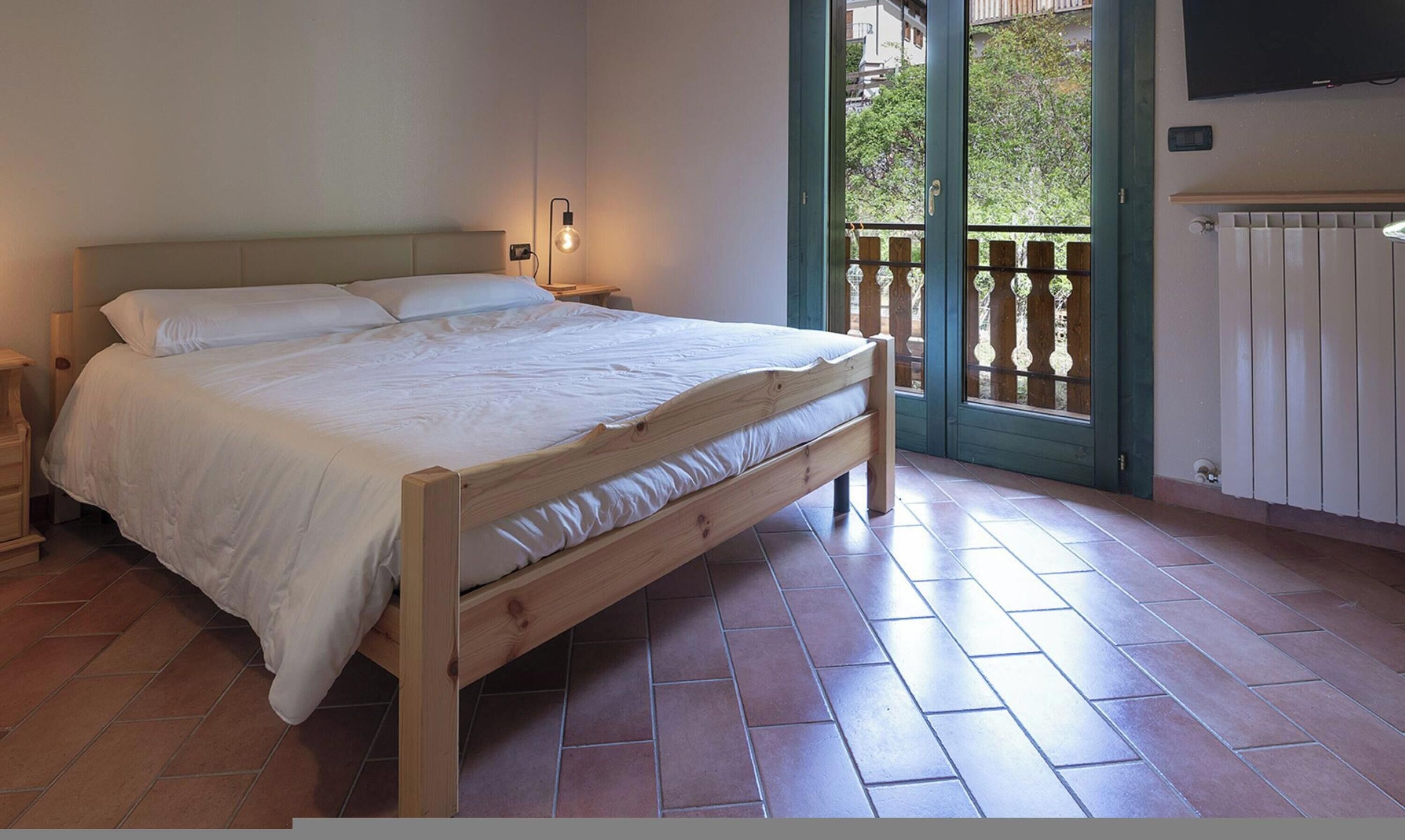 A bedroom with a wooden bed, a nightstand, a wall-mounted light, a radiator, and a balcony with a wooden railing.