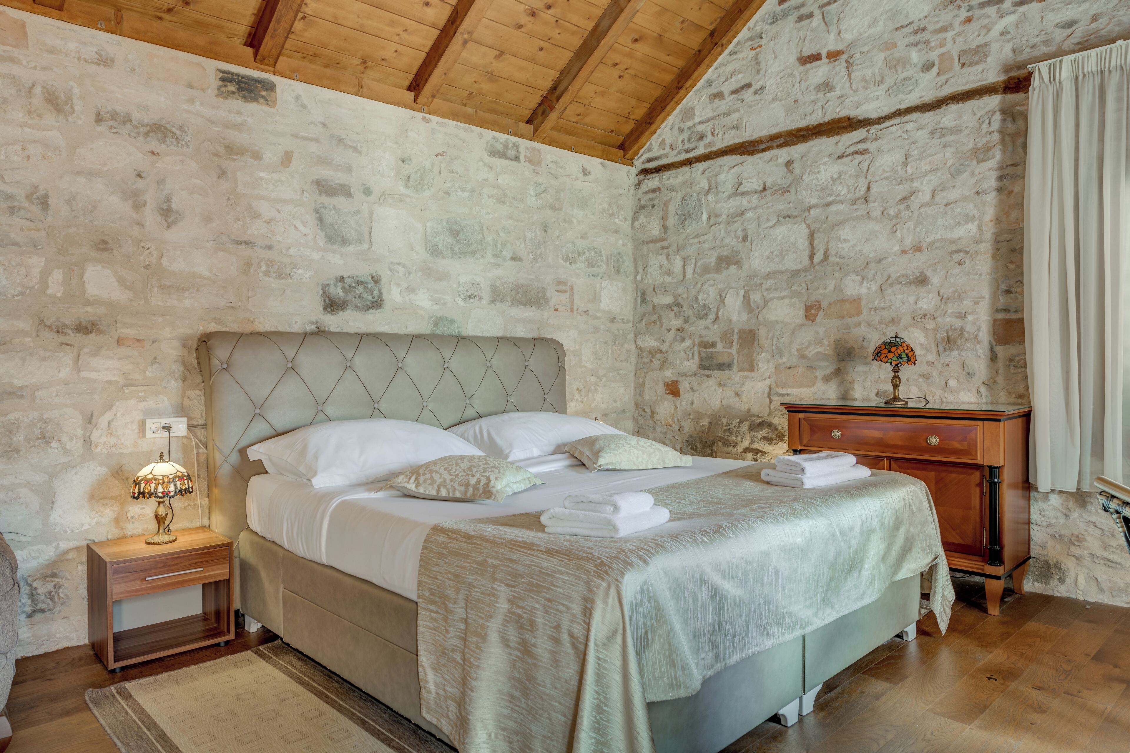 A bedroom with a stone wall, a wooden headboard, a bedside table with a lamp, and a nightstand.