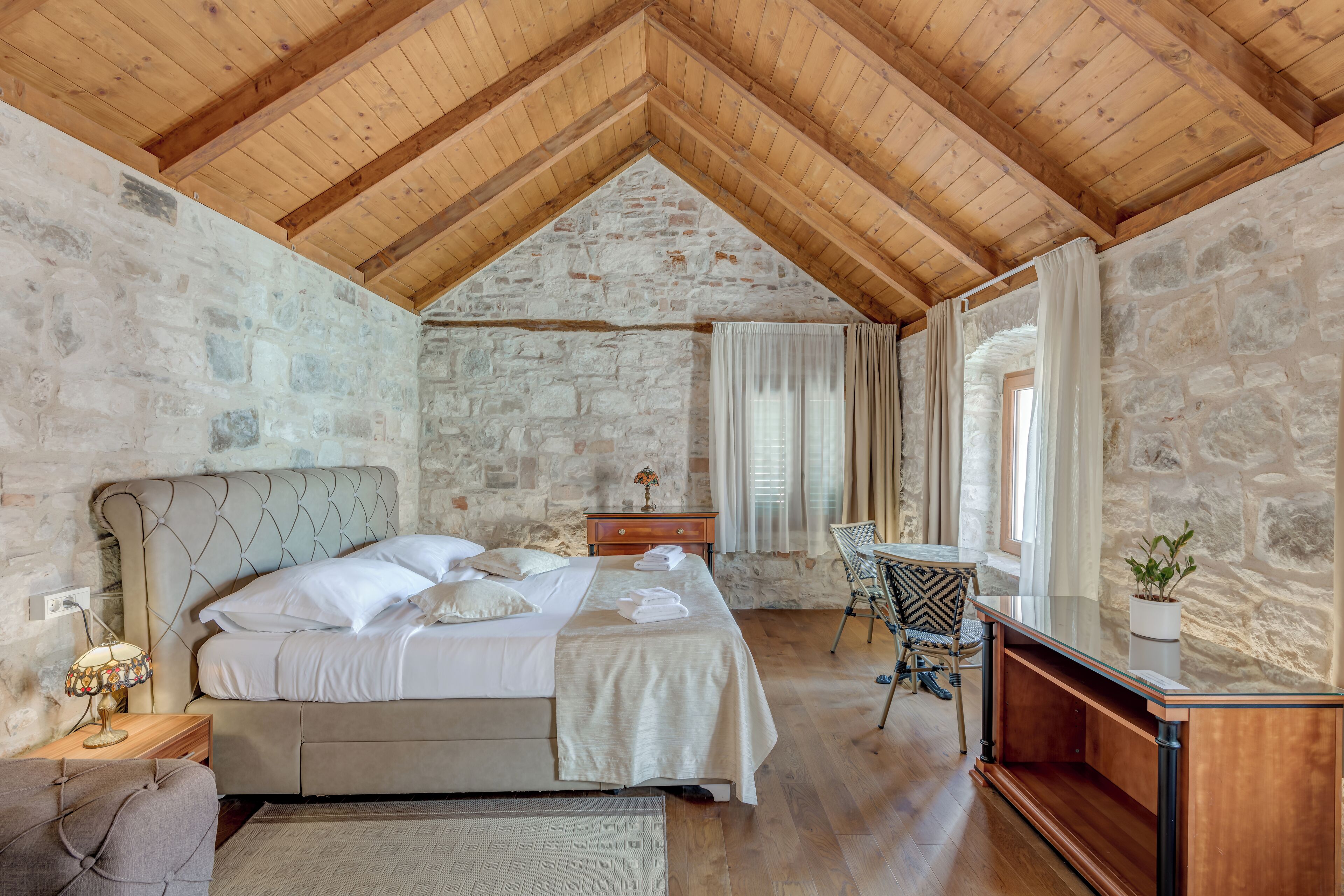 A bedroom with a stone wall, wooden ceiling beams, a bed, a desk, and a chair.