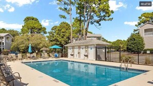 A rectangular swimming pool with lounge chairs and umbrellas, surrounded by trees and a small building with a roof.