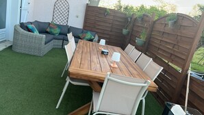 Un patio exterior con una mesa de madera y sillas blancas, rodeado por una valla de madera y plantas en macetas.