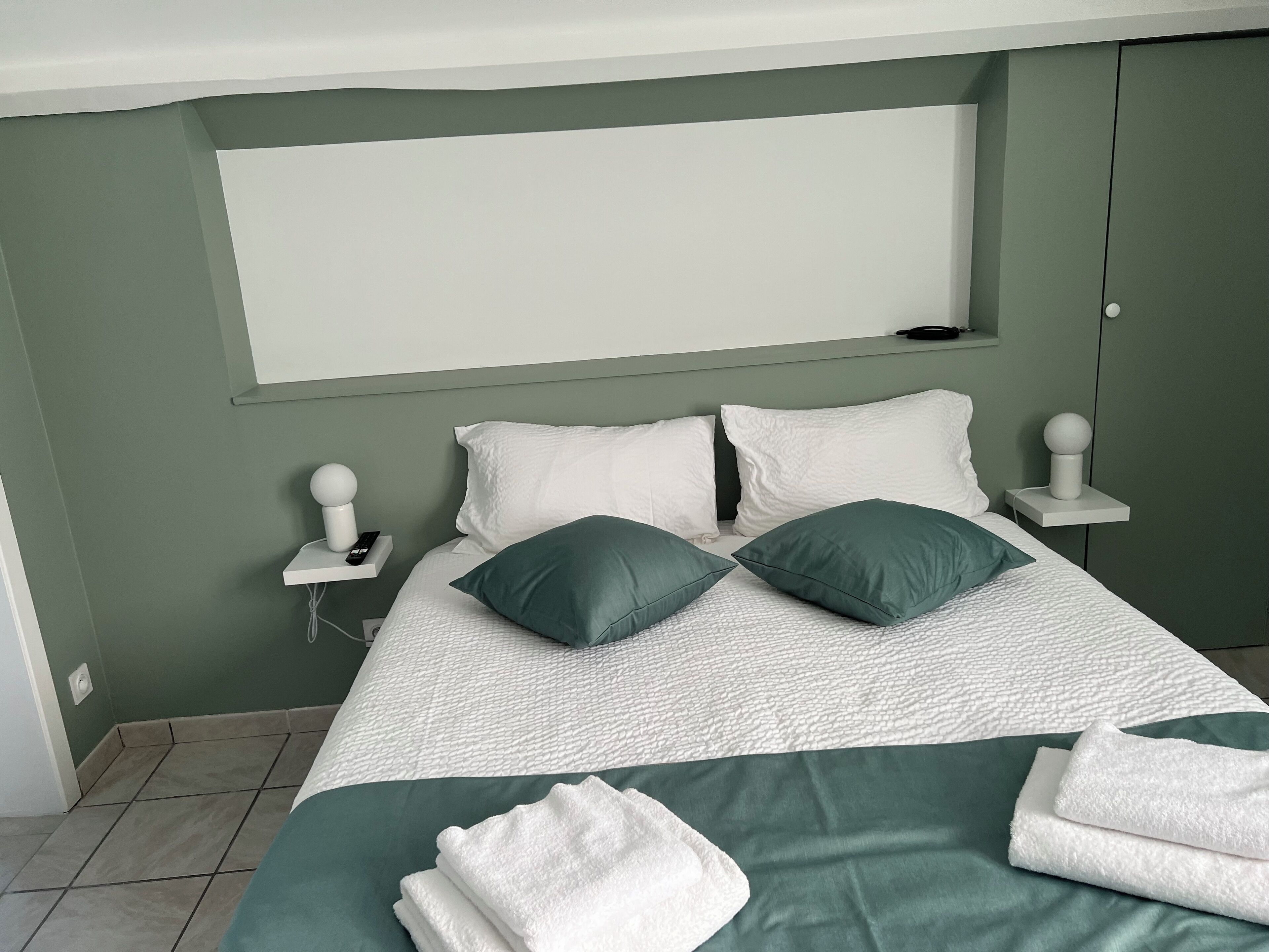 A neatly made bed with white and green bedding, two bedside tables with lamps, and a plain wall.
