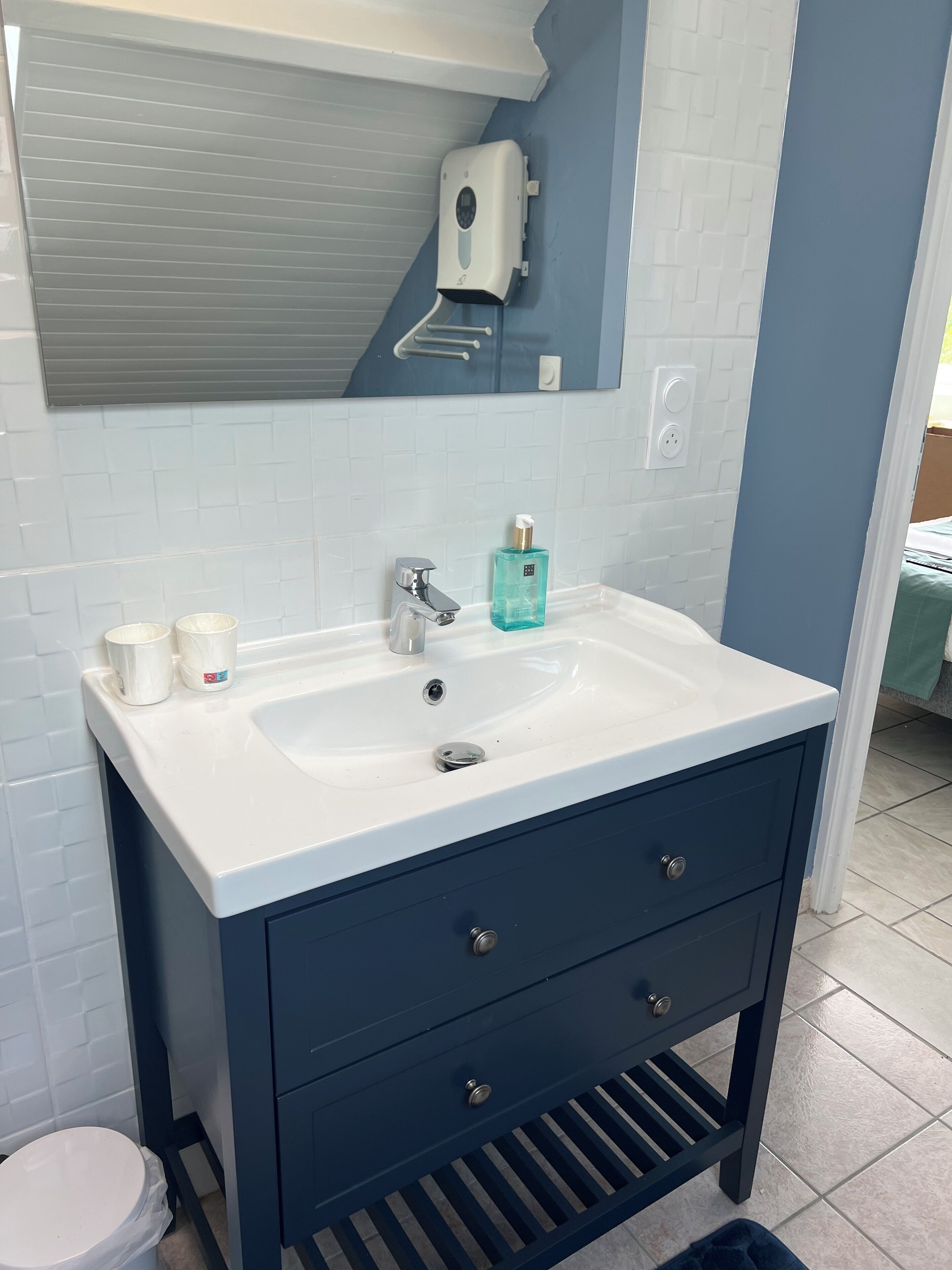Ein Badezimmer mit einem weißen Waschbecken, einem dunkelblauen Schrank und einem Spiegel über dem Waschbecken. Auf der Waschbeckenablage stehen zwei weiße Tassen und ein blauer Seifenspender.