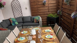Una mesa de comedor al aire libre, impecablemente dispuesta, con varios juegos de cubertería, vasos y un llamativo muro decorativo al fondo.