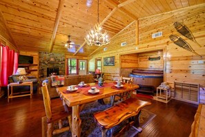 A wooden cabin interior with a dining area, a fireplace, a stone wall, and a hot tub. - Lost By The Pond Cabin | Hochatown Cabin with Fishing Pond, Hot Tub, Fireplace & Pets (Hochatown)