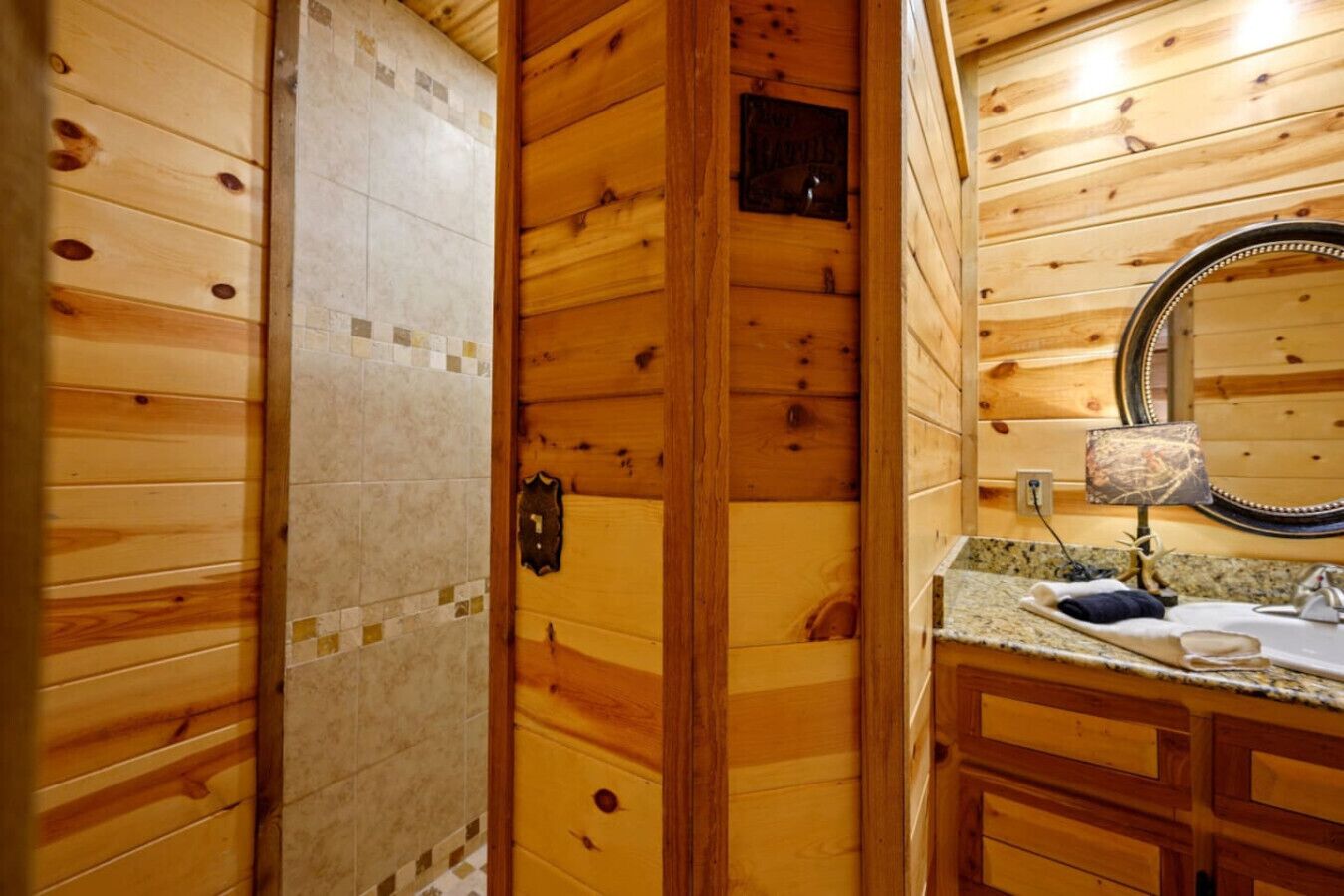 Een houten badkamer met een betegelde douche, een houten deur met nummerplaat, een wastafel met granieten aanrechtblad en een ronde spiegel.
