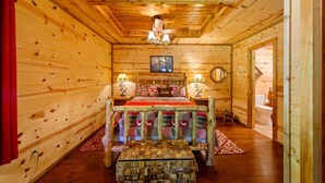 A wooden cabin bedroom with a bed, bedside lamps, a chandelier, and a red curtain. - Lost By The Pond Cabin | Hochatown Cabin with Fishing Pond, Hot Tub, Fireplace & Pets (Hochatown)