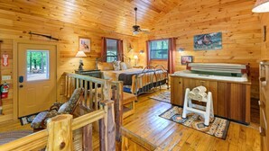 A wooden cabin interior with a bed, a hot tub, a fireplace, and a window with a view of trees. - Lost In The West Cabin | Hochatown Cabin with Fishing Pond, Hot Tub, Fireplace & Pets (Hochatown)