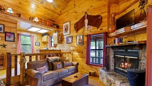 A cozy cabin interior with a stone fireplace, wooden walls, a sofa, and a television. - Lost In The West Cabin | Hochatown Cabin with Fishing Pond, Hot Tub, Fireplace & Pets (Hochatown)