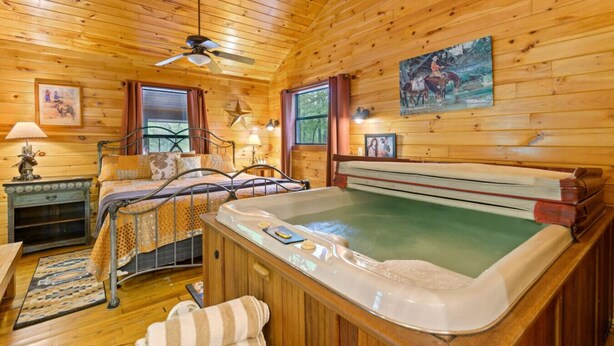 A wooden cabin room with a bed, a hot tub, a ceiling fan, and a painting on the wall. - Lost In The West Cabin | Hochatown Cabin with Fishing Pond, Hot Tub, Fireplace & Pets (Hochatown)