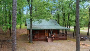 A cabin with a porch, surrounded by trees. - Lost In The West Cabin | Hochatown Cabin with Fishing Pond, Hot Tub, Fireplace & Pets (Hochatown)