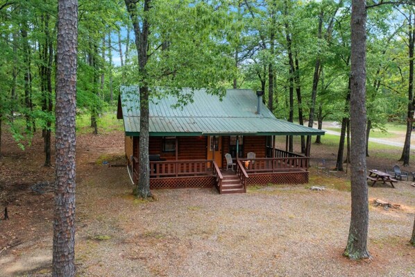 A cabin with a porch, surrounded by trees. - Lost In The West Cabin | Hochatown Cabin with Fishing Pond, Hot Tub, Fireplace & Pets (Hochatown)