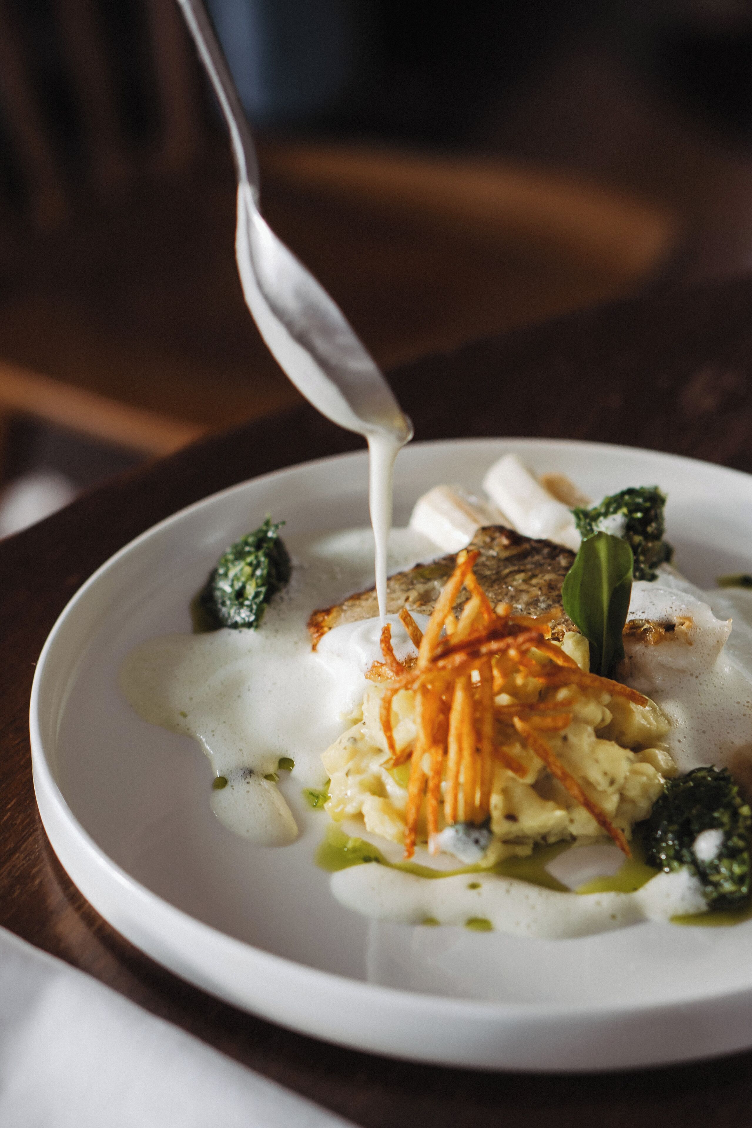 A white plate with a variety of food items including a piece of fish, vegetables, and a creamy sauce being poured over it.