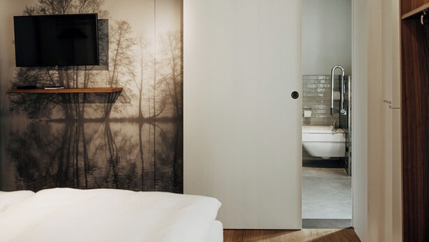 A bedroom with a bed, a TV on a stand, and a door leading to a bathroom with a shower and toilet. - Bootshaus Amberg  (Amberg)