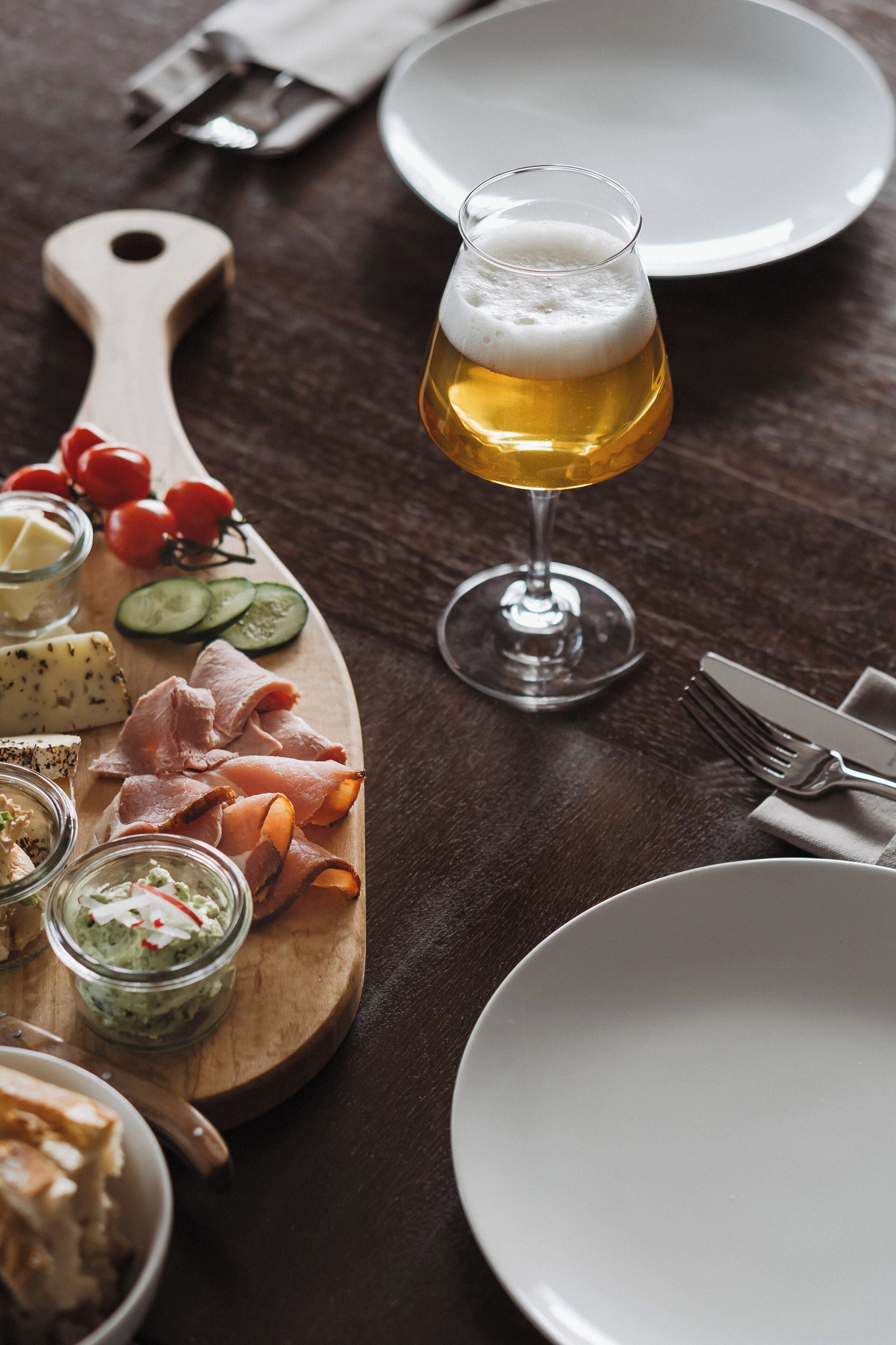 A wooden board with various cheeses, meats, and vegetables, a glass of beer, and a knife with a wooden handle on a dark wooden table.