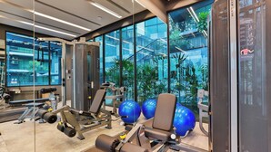 A modern gym with various exercise equipment, large windows, and a view of a building with outdoor plants. - B305 - 1 Bedroom Serviced Apartment Pool View near Ao Nang Beach (Tambon Ao Nang)