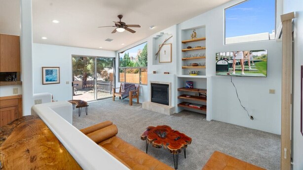 A modern living room with a fireplace, a television mounted on the wall, and a large window providing a view of the outdoors.