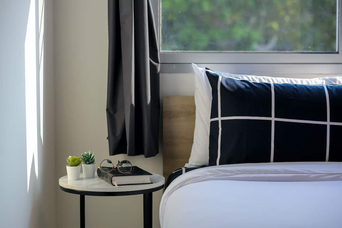 A neatly made bed with a white and navy pillow, a small bedside table with a book and two potted plants, and a window with a view of greenery.