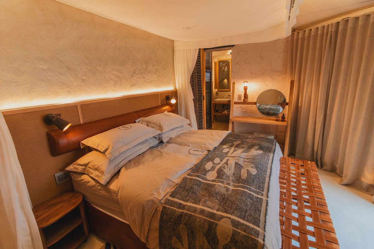 Een hotelkamer met een net opgemaakt bed, een houten nachtkastje, een bijzettafeltje, een spiegel en uitzicht op de gang.