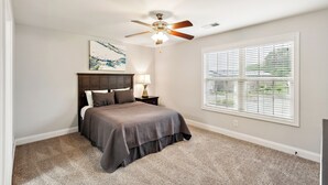 A bedroom with a bed, a ceiling fan, a nightstand with a lamp, a window with blinds, and a painting on the wall.