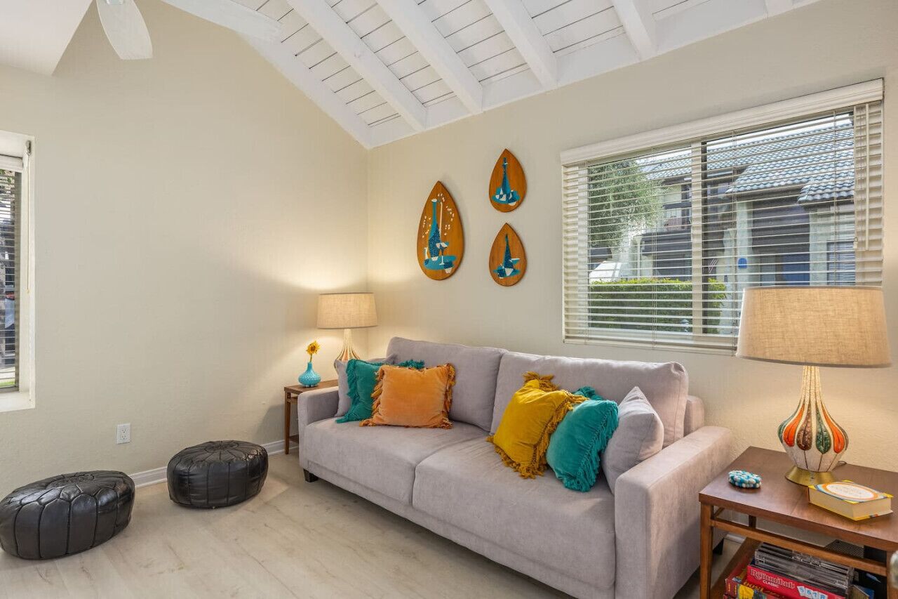 Un salon avec un canapé gris, des poufs noirs, une petite table avec une lampe et une fenêtre avec des stores.