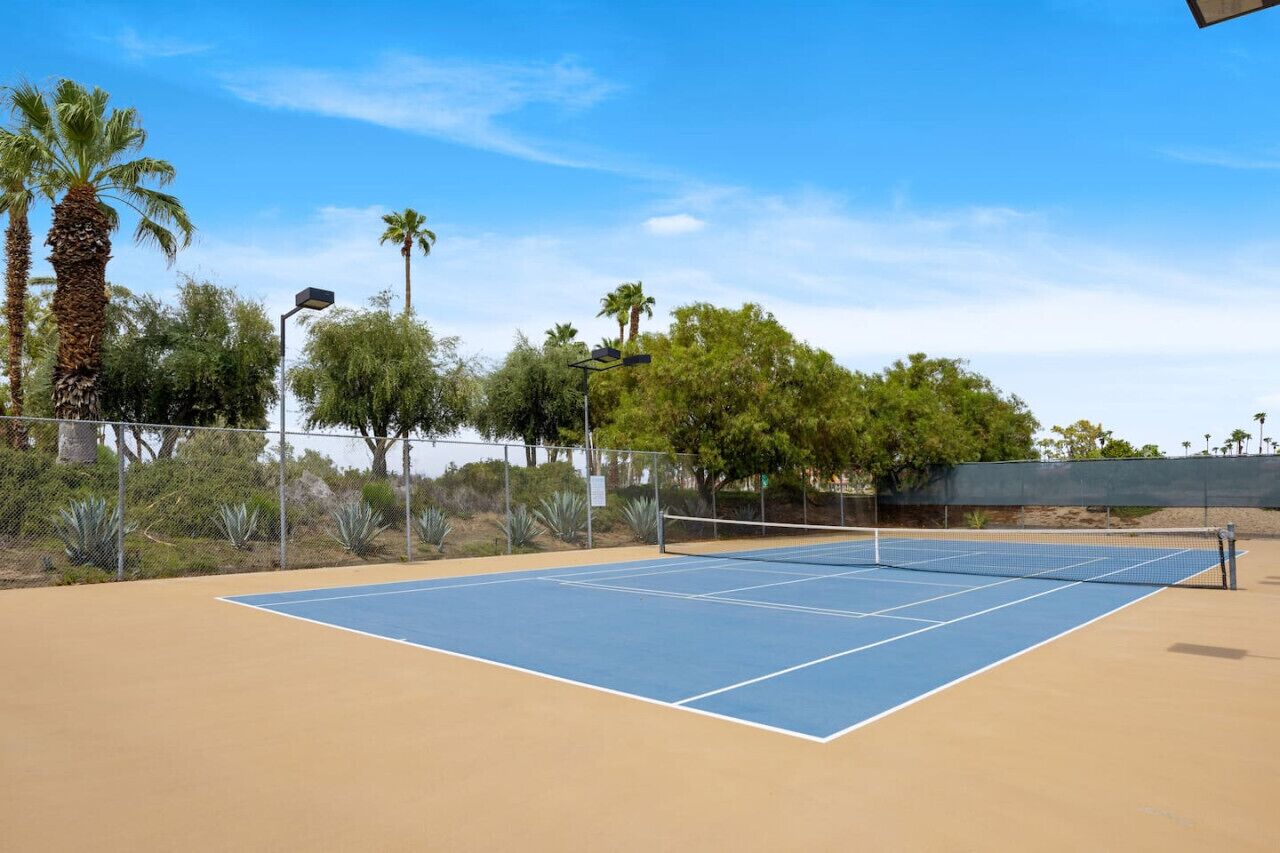 Una cancha de tenis al aire libre con superficie azul, rodeada por una cerca, palmeras y otra vegetación.