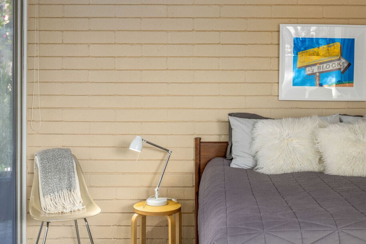 Un dormitorio con pared de ladrillo, una cama con colcha gris, almohadas mullidas, una mesita redonda con lámpara y una silla con manta gris.
