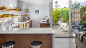 A modern kitchen with wooden shelves, a large island, and a view of a backyard with outdoor furniture. - MidMod Racquet Club Villa (Palm Springs)