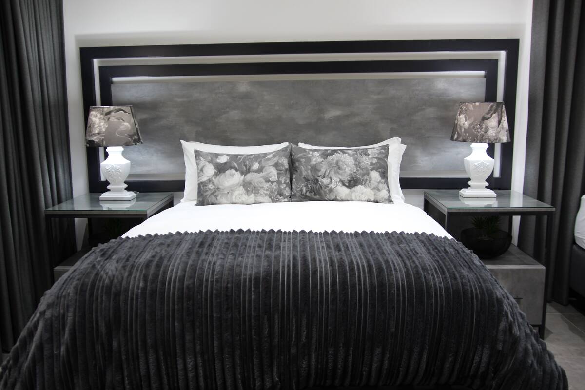 Un dormitorio con una paleta de colores blanco y negro, que incluye una cama grande con manta negra, dos mesitas de noche con lámparas y un cabecero gris.