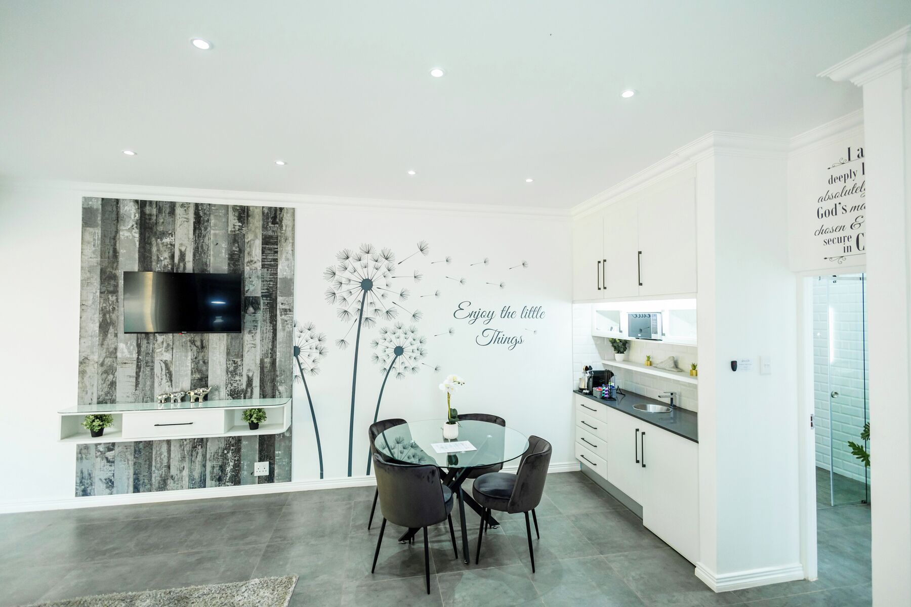A modern kitchen with white cabinets, a glass dining table, and a wall-mounted TV.