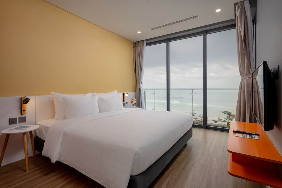 Une chambre d’hôtel moderne avec un grand lit, une petite table de chevet, une télévision et une vue sur l’océan.