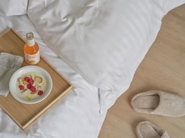 A bed with white linens, a wooden tray with a bowl of cereal and a bottle of orange juice, and a pair of slippers.