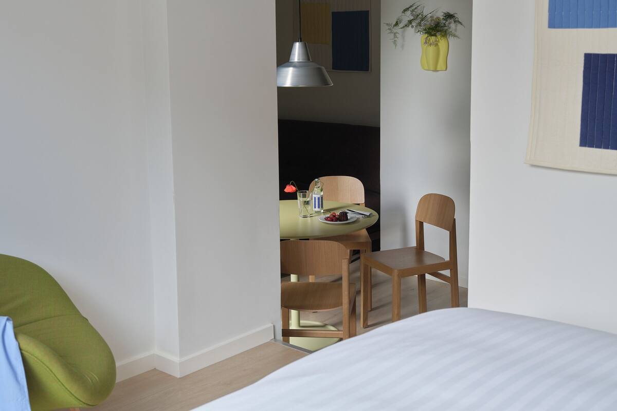 Quarto de hotel com uma cama, uma mesa de jantar com cadeiras, uma planta pendente e um painel com obras de arte emolduradas.