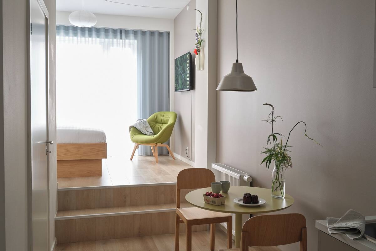 Quarto moderno e bem iluminado, com área de refeições, uma cadeira verde, uma mesa com uma tigela de frutas e um radiador.