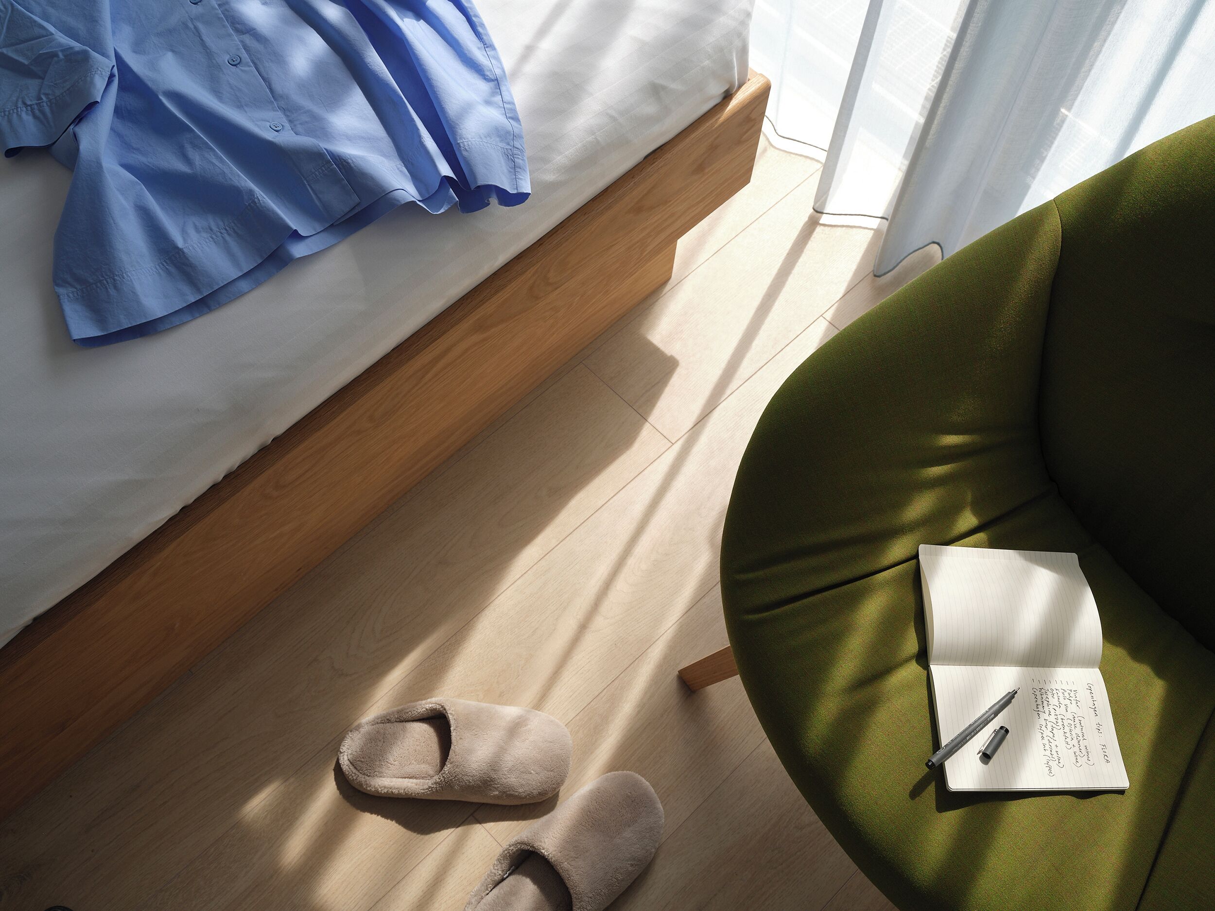 Ein Schlafzimmer mit einem Bett, einem grünen Sessel, Hausschuhen und einem Notizbuch mit Stift.