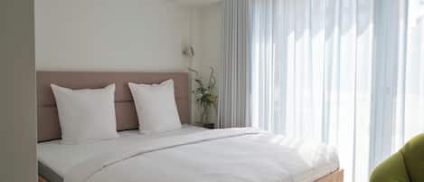 Un dormitorio moderno con estructura de cama de madera, ropa de cama blanca y cortinas translúcidas que dejan entrar la luz natural.