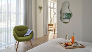 Habitación de hotel con cama, una silla verde, bandeja de desayuno con fruta y jugo, y vista al exterior a través de amplias ventanas.