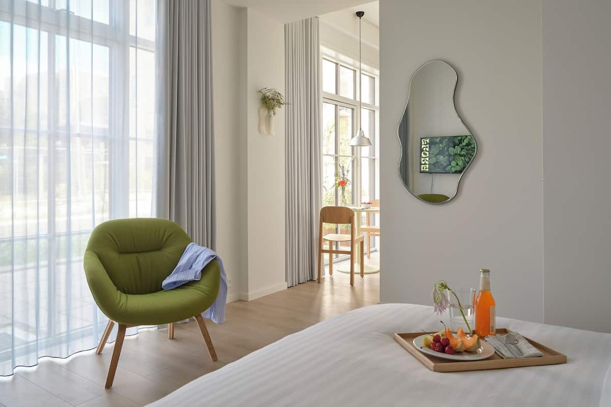 Quarto de hotel com cama, uma cadeira verde, bandeja de café da manhã com frutas e suco, e vista para o exterior através de grandes janelas.